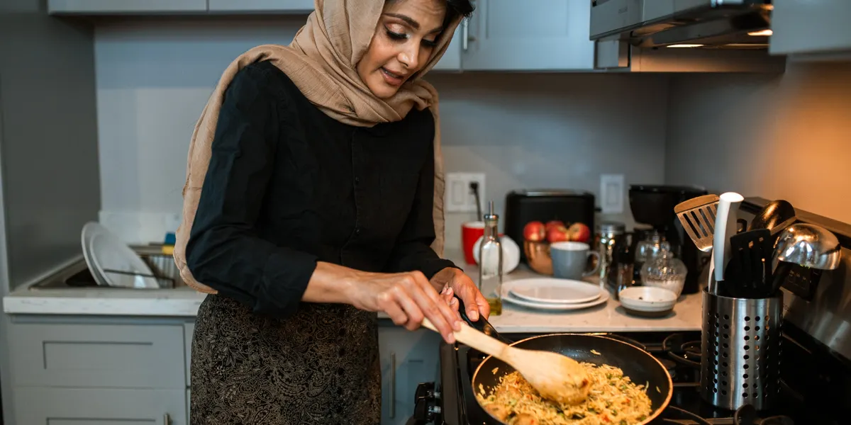 A woman in a headscarf stirring a pan of rice and vegetables with a wooden spoon on a home cooktop