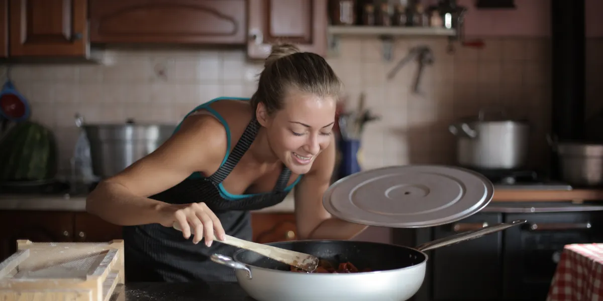 A woman at a home stove lifting the lid off a frying pan and stirring with a wooden spoon